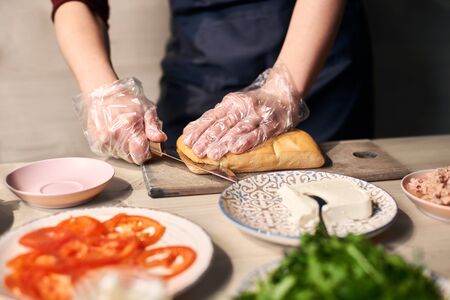 Focus on hand cutting with knife long bread roll on cutting board for preparing grilled panini sandwich. Ingredients on blurred foreground: tomato slices, tuna, butter, salad on plate at wooden table.の写真素材