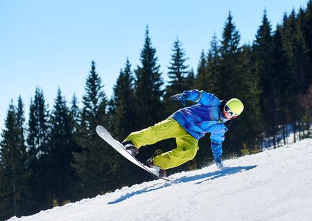 Snowboarder man in goggles and helmet riding snowboard fast down steep snowy mountain slope on background of blue sky and spruce trees on sunny winter day. Extreme sport and recreation concept.の写真素材
