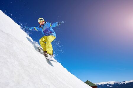 Guy snowboarding down the mountain slope. Portrait of young man snowboarding in winter against blue sky. Clear weather and sky, prepared snowの写真素材