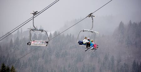 Bukovel, Ukraine - December 09, 2018: family three persons skiers with skis on their feet sitting on cable chairlift carrying up over dark coniferous forest covered with dense fog in winter mountainsのeditorial素材