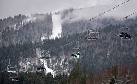 Bukovel, Ukraine - December 09, 2018: pairs of skiers with skis on their feet sitting on cable chairlift carrying up over slope and dark mountain coniferous forest with fog in winter mountainsのeditorial素材