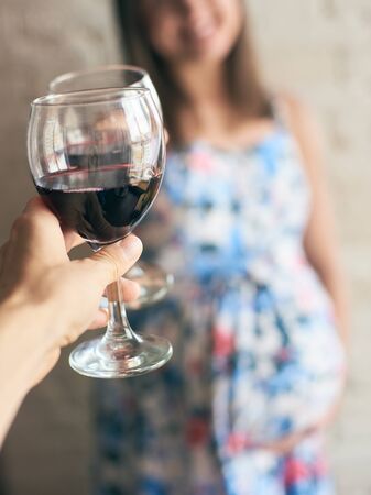 Pregnant woman keeping glass of wine, cheering with friend and laughing. Future mother drinking alcohol while expecting little baby. Concept of unhealthy lifestyle and harmfulness.の写真素材