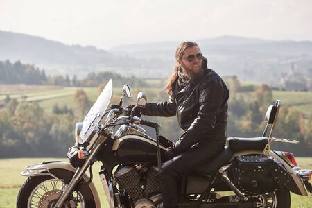 Happy handsome bearded biker with long hair in black leather jacket and sunglasses sitting on cruiser motorcycle, on blurred background of green peaceful rural landscape and light foggy sky.の写真素材