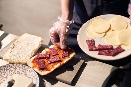 Chef hands holding white plate with slices of cheese and sausage. Cook in gloves putting pieces of sausage on bread greased with red sauce. Butter in plate with ornament, cutting board on background.の写真素材