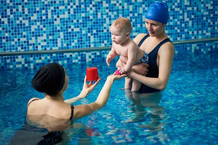 Early development sport class for infant. Active educational games over water in paddling pool with parents and specialist. Cute baby boy interested in communication with young female swimming trainerの写真素材