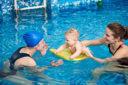 Adorable baby enjoying swimming on floating board in pool with his mother and trainer helping. Side view. Interesting game and useful pastime for children. Newborn baby at sportive active familyの写真素材
