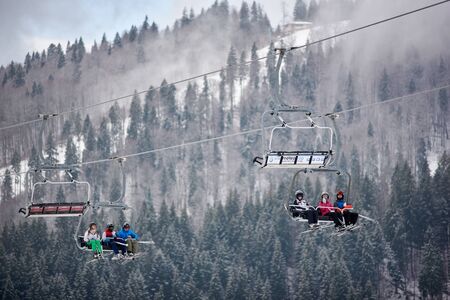 Bukovel, Ukraine - December 09, 2018: skiers and snowboarders sitting carrying high up on cable chairlift in one direction over snow-covered slopes in Carpathians mountains in foggy gray dayのeditorial素材