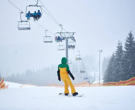 Back view of unrecognizable snowboarder sliding on high slope protected by net under chairlift carrying people up to mountain top for skiing. Foggy winter wooded mountain landscape on backgroundの写真素材