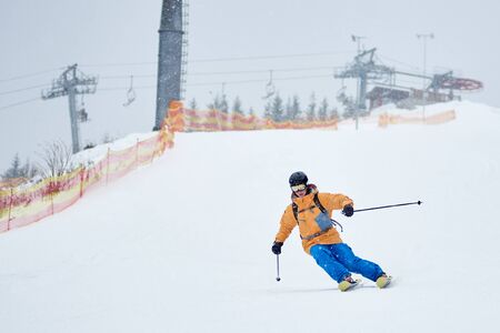 Proficient young skier skiing downhill from high snow-covered slope with net on edge. Winter sports activities in dense snowfall at ski resort. Mastery skiing skills. Lift station on mountain top.の写真素材