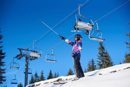 Female skier smiling standing on mountain slope near ski lift hoist. Woman in ski goggles showing with ski poles something far away on sunny winter day. Active lifestyle at resort. View from low angleの写真素材