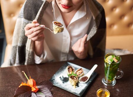 Elegant girl in white blouse eating tasty sweet sushi roll with chocolate sauce decorated with icing sugar. Woman with red lips covered in blanket relaxing with dessert and mint tea with honey.の写真素材