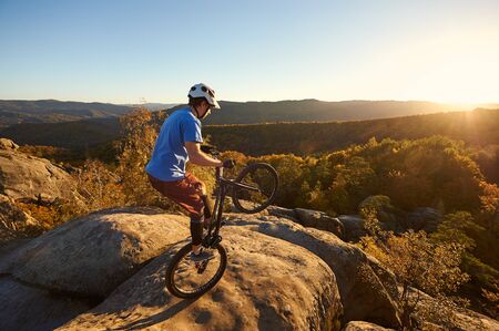 Fearless cyclist balancing on back wheel on trial bike. Sportsman biker making acrobatic stunt on the edge of big boulder on the top of mountain at sunset. Concept of extreme sport active lifestyleの写真素材