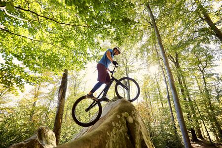 Male professional cyclist balancing on trial bicycle, making acrobatic trick on big boulder in the forest outdoor on summer sunny day. Concept of extreme sport active lifestyleの写真素材