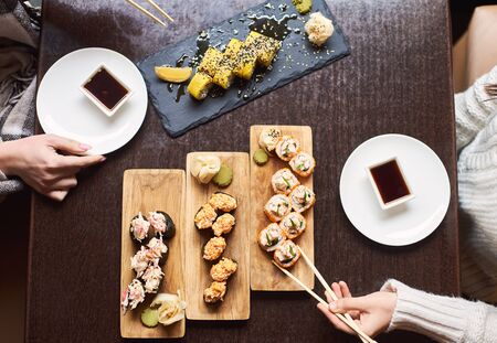 Top view of female hands in oriental restaurant. Women enjoying sushi set with soy sauce, wasabi and marinated ginger. Friends eating popular Japanese dish from rice and seafood with chopsticks.の写真素材