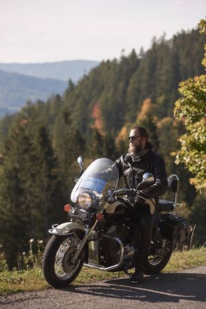 Handsome bearded biker in black leather jacket and sunglasses resting on cruiser motorcycle on country roadside, on blurred background of foggy green hills covered with dense spruce forest.の写真素材