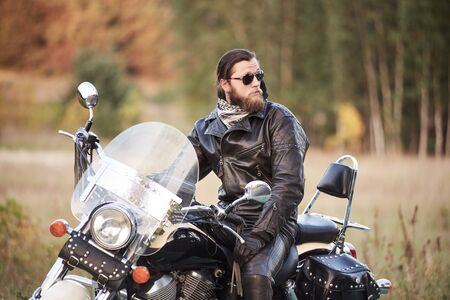 Young bearded motorcyclist in black leather clothing and dark sunglasses sitting on black shiny modern motorbike, on blurred vintage sunny bokeh background of golden-green trees foliage on sunny day.の写真素材