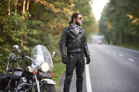 Profile bearded athletic long-haired motorcyclist in black leather outfit and dark sunglasses standing at shiny cruiser powerful motorbike, on blurred background of hilly asphalt road and green trees.の写真素材