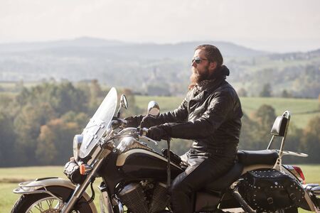 Side view of handsome bearded biker with long hair in black leather jacket and sunglasses sitting on cruiser motorcycle, on blurred background of green peaceful rural landscape and light foggy sky.の写真素材