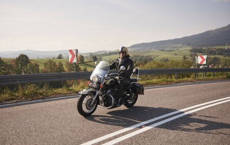 Bearded biker in helmet, sunglasses and black leather clothing riding cruiser motorcycle along sharp turn of empty road on bright summer day on misty background of rural landscape and distant hills.の写真素材