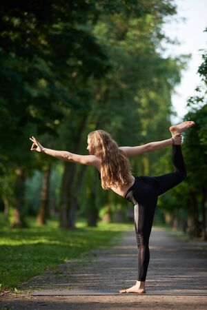 Woman with long hair in black sports suit standing in pose of king of dance, Asan Natarajasana, against greenery park under sunny rays. Morning training outdoors at summer. Healthy lifestyle conceptの写真素材