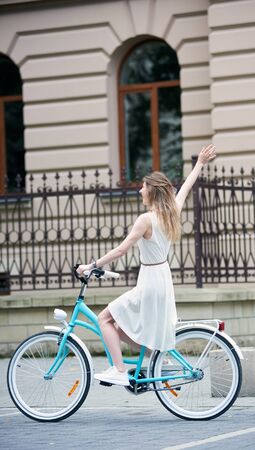 Young woman in light white dress and white sneakers is riding a blue bike on the street in the city with one hand up, full length, beautiful building with fence on background, side viewの写真素材