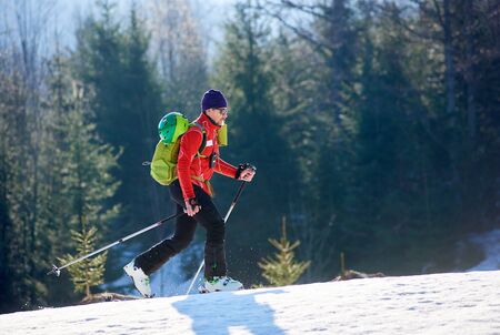 Happy sportive male skier tourist with backpack in sunglasses walking on skis uphill in deep snow on background of winter wood on sunny cold day. Holiday touring, sport and exploration concept.の写真素材