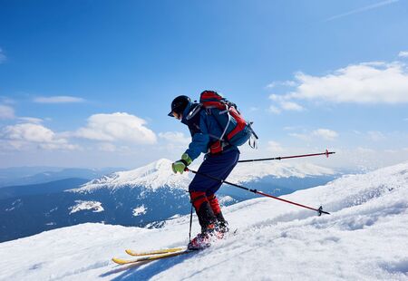 Sportsman skier in bright clothing, helmet and goggles with backpack riding down steep snowy slope on copy space background of blue sky and mountain landscape. Winter holidays, extreme sport concept.の写真素材