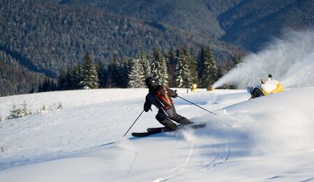 Back view of man skiing on prepared hill with fresh new powder snow. Artificial snowfall in sunny day. Electric snow gun machine. Ski lesson in skiing school. Scenic wooded mountains on background.の写真素材