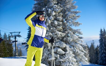 Female skier in goggles shielding from sun with her hand and looking for someone. Smiling woman coming up on slope top with ski lift. Skier before freeride-descending. Mountain view on background.の写真素材