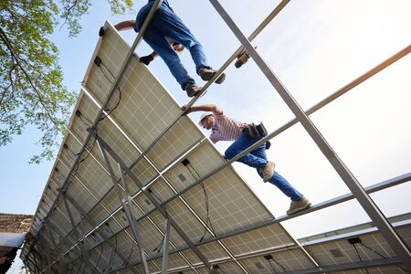 Bottom view from inside of stand-alone exterior solar panel system with two engineers working on high steel platform on bright clear blue summer sky background. Ecological energy production concept.の写真素材