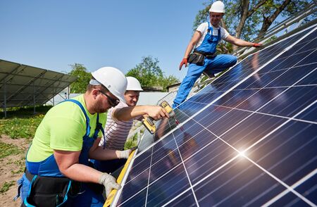 Team of workers taking measurement and connecting solar photo voltaic panels to high steel platform under blue sky. Stand-alone solar panel system installation, efficiency and professionalism concept.の写真素材