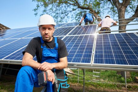 Portrait of successful engineer technician with electrical screwdriver in front of almost finished high exterior solar panel photo voltaic system, with team of workers on high platform.の写真素材