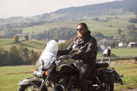 Side view of handsome bearded motorcyclist with long hair in black leather jacket and sunglasses, sitting on modern motorcycle on blurred background of green peaceful rural landscape.の写真素材