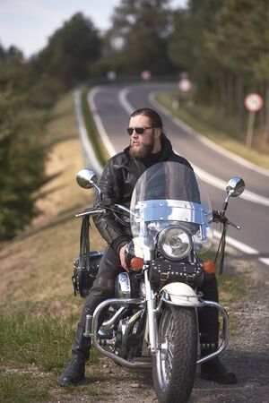 Handsome bearded biker with long hair in leather jacket and sunglasses sitting on cruiser motorcycle on roadside, on blurred background of empty twisty road with white marking on sunny summer day.の写真素材