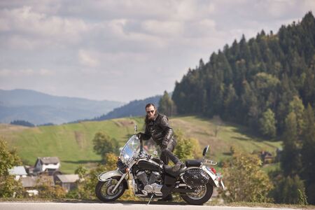 Side view of handsome bearded biker in black leather jacket and sunglasses sitting on cruiser motorcycle on roadside on blurred background of foggy spruce forest, green hills and bright cloudy sky.の写真素材
