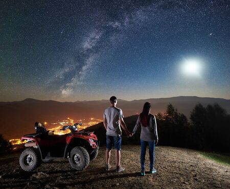 Back view of young couple tourists with atv quad motorbike on the top of mountain. Man and woman holding hands, enjoying night sky full of stars, full moon, Milky way, luminous city on backgroundの写真素材