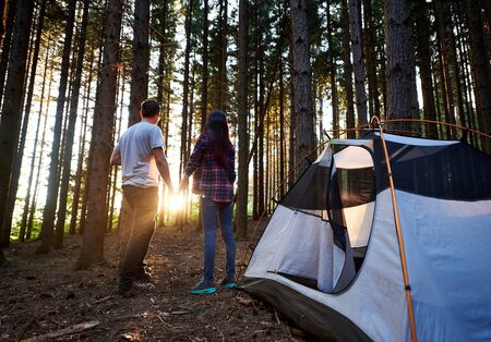 Back view of man and woman travellers holding hands, standing near white tourist tent. Romantic couple enjoying beautiful view of setting sun. Evening camping at sunset in the forest.の写真素材