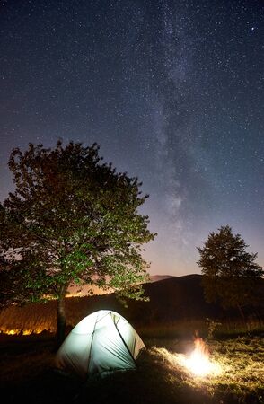 Tourist camping near big tree at night. Illuminated tent and bonfire under amazing night sky full of stars and Milky way. On the background beautiful starry sky and mountainsの写真素材