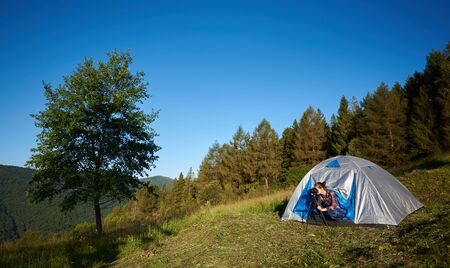 Summer morning camping in the mountains. Young woman hiker sitting in tourist tent, taking photo, using photo camera and tripod. On background blue sky, forest and big tree.の写真素材
