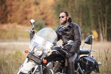 Handsome bearded motorcyclist in black leather clothing and dark sunglasses sitting on black shiny cruiser bike, on blurred vintage sunny bokeh background of golden-green trees foliage in the evening.の写真素材
