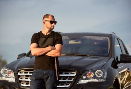 Portrait of a successful self-confident man standing in front of his black car with crossed hands, looking away, wearing dark sunglasses, black t-shirt and jeans, close-up, copy spaceの写真素材