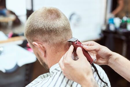 Dark blonde man at barber shop is having his nape shaved with a trimmer, hairdresser is holding a red tool, stripe cape onの写真素材