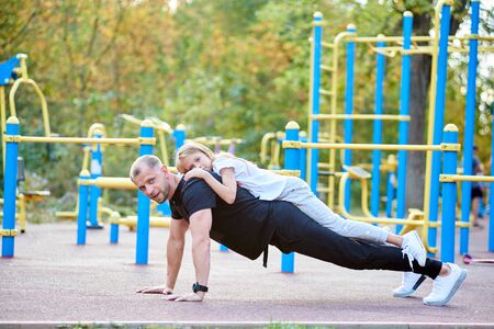 Father is doing push ups on the ground outdoors with his daughter on his back, looking in the camera, healthy lifestyle with family, workout, side viewの写真素材