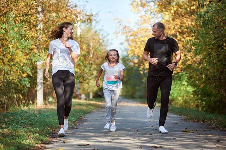Front view of mother, father and daughter wearing sportswear are jogging in autumn park alley, active family, warm and sunny weatherの写真素材