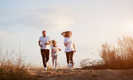 Front view of young and beautiful family of three jogging with their dog outside the city on the village road on the setting sunの写真素材