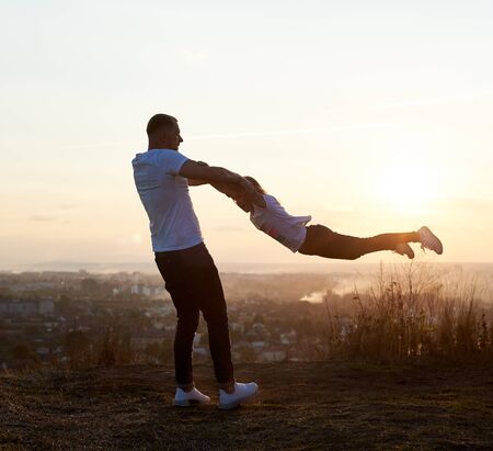Silhouette of a father swinging his daughter by the arms on the hill on the setting sun in the suburbs, beautiful horizon on the background, side viewの写真素材