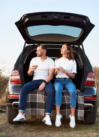 Man and woman are sitting in the car trunk on a check blanket, holding glasses of white wine, wearing similar clothes, looking awayの写真素材