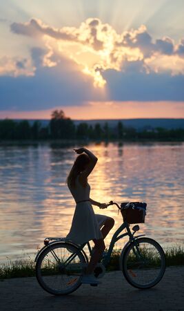 Lady in a light dress rides a vintage bicycle with a basket near the river against the backdrop of a bewitching sunset. The rays of the sun at sunset break through the cloudsの写真素材
