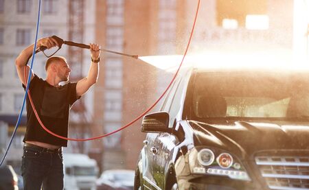 Close-up view of a man cleaning his black car outdoors, water is splashing over the top of the car shining on the sun.の写真素材
