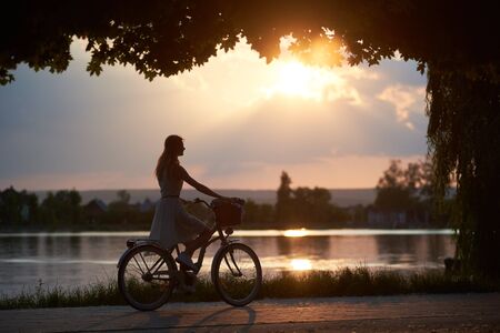 Young woman rides a vintage bicycle with a basket on the road near the river on a sunset. Girl enjoys the magical view of the sun's rays of the falling sun at the end of the dayの写真素材